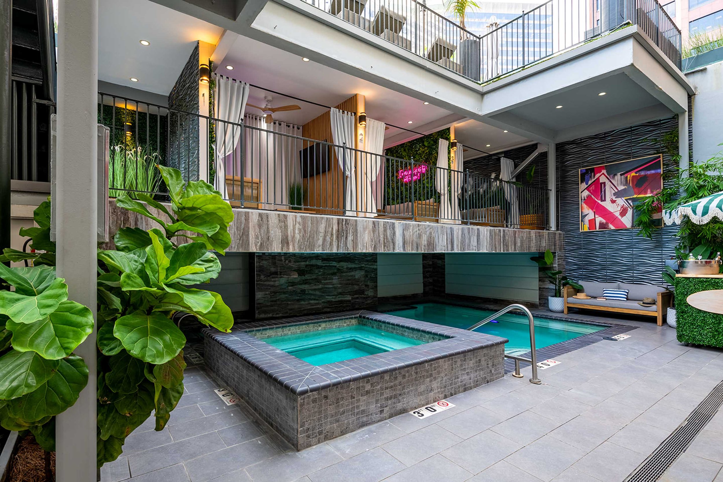 Indoor pool area with hot tubs, a seating area with a couch and a decorative wall painting, plants, and a balcony above.