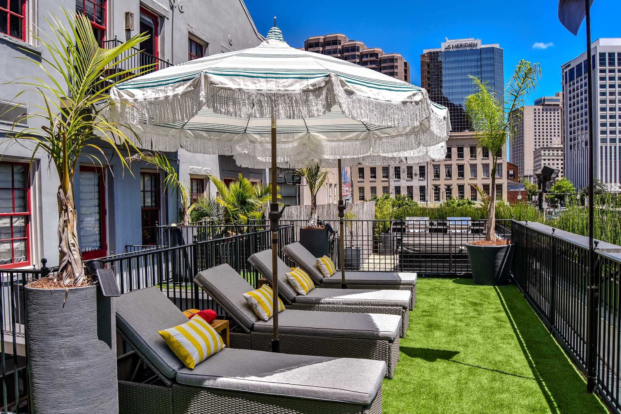 Outdoor rooftop patio with four lounge chairs, a large white striped umbrella, and potted palm trees, surrounded by a black railing, with city buildings in the background and a clear blue sky.